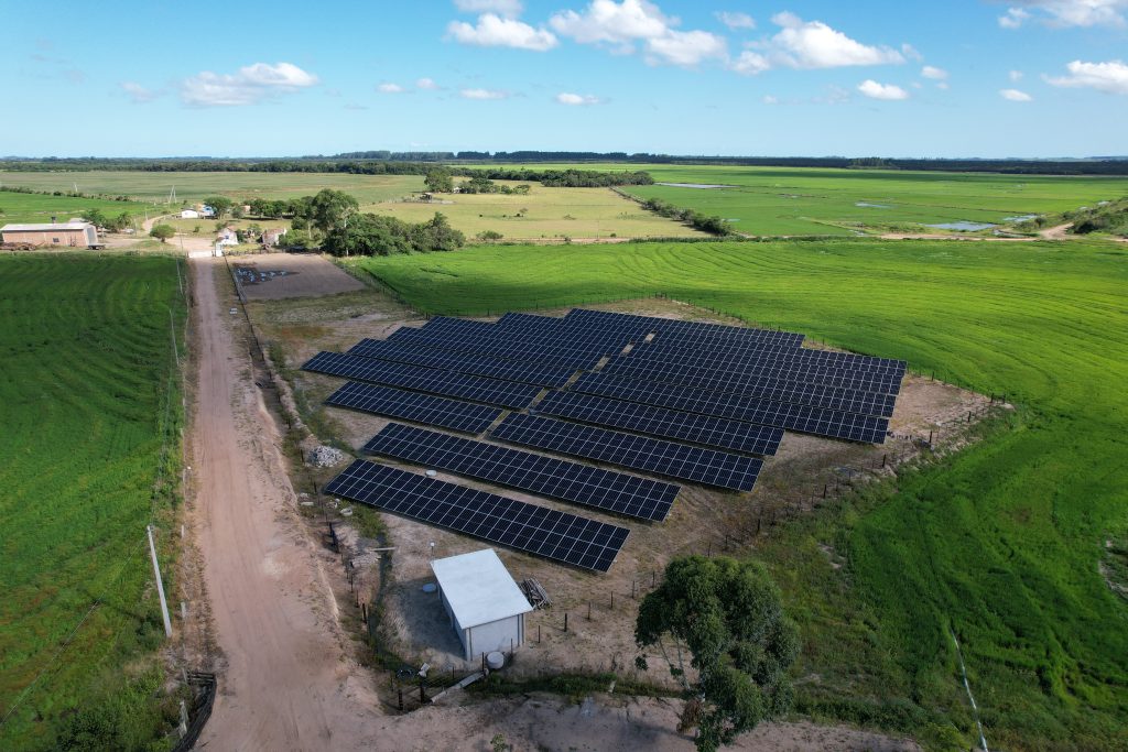 Vista aérea de uma usina solar fotovoltaica instalada em área rural, cercada por campos verdes e céu azul.